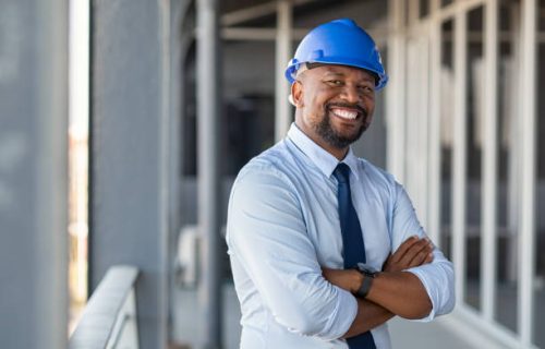 Portrait of african american man architect at building site with folded arms looking at camera. Confident construction manager in formal clothing wearing blue hardhat. Successful mature civil engineer at construction site with copy space.